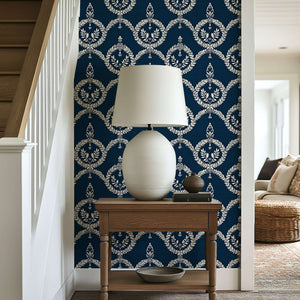 Dark navy wallpaper with ivory laurel wreath patterns, enhancing an elegant and traditional look in a hallway with a lamp and table.