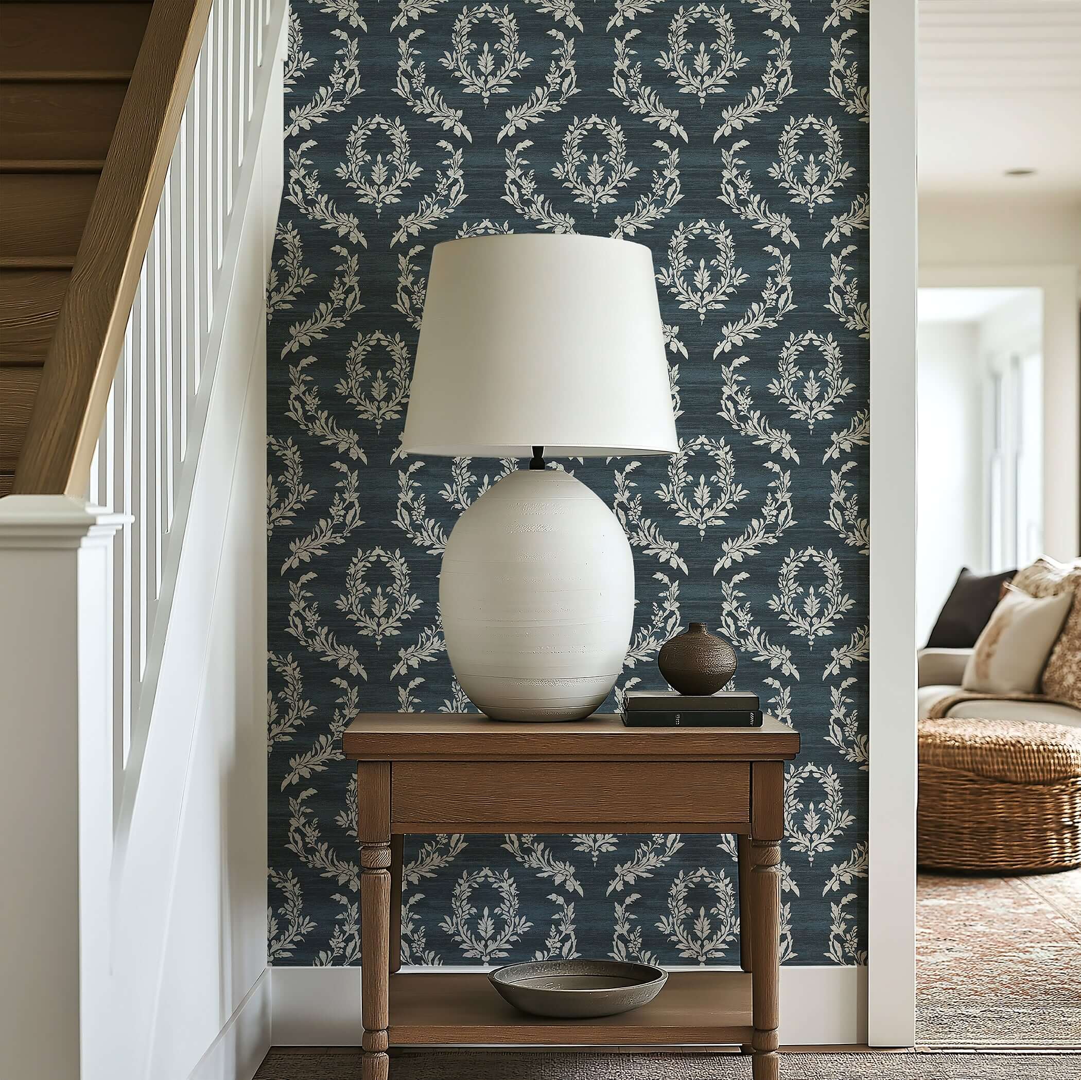 Midnight Laurel Wallpaper featuring white laurel wreaths on dark blue backdrop in a stylish room with a wooden table and lamp.