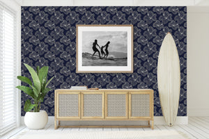 Island Nights Floral Wallpaper with hibiscus on navy background, enhancing a coastal living room setting with surfboard and plant decor.