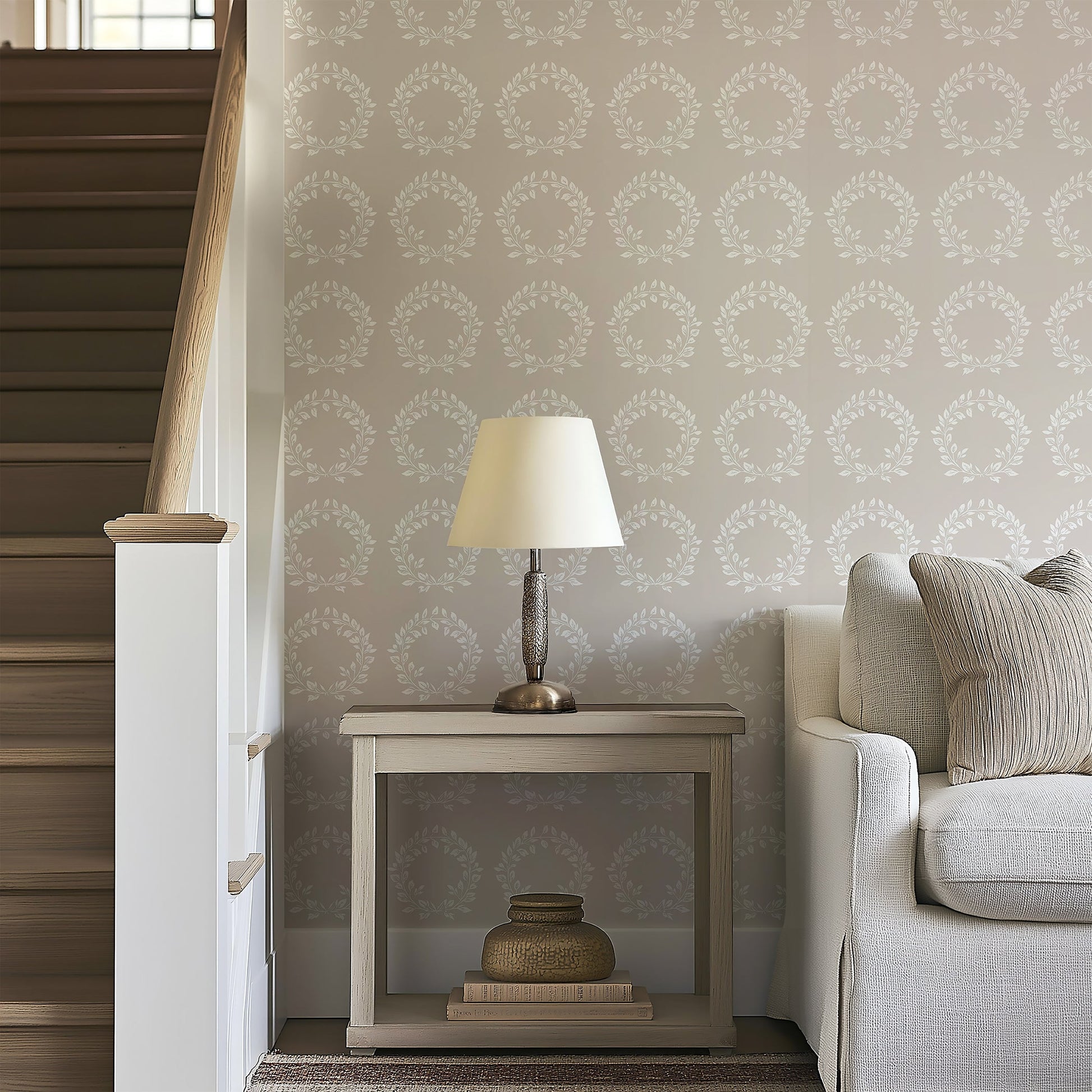 Living room with Traditional Wreath Harmony wallpaper featuring white laurel wreaths on taupe, alongside a sofa and table lamp.