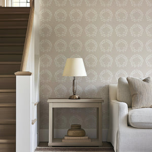 Living room with Traditional Wreath Harmony wallpaper featuring white laurel wreaths on taupe, alongside a sofa and table lamp.
