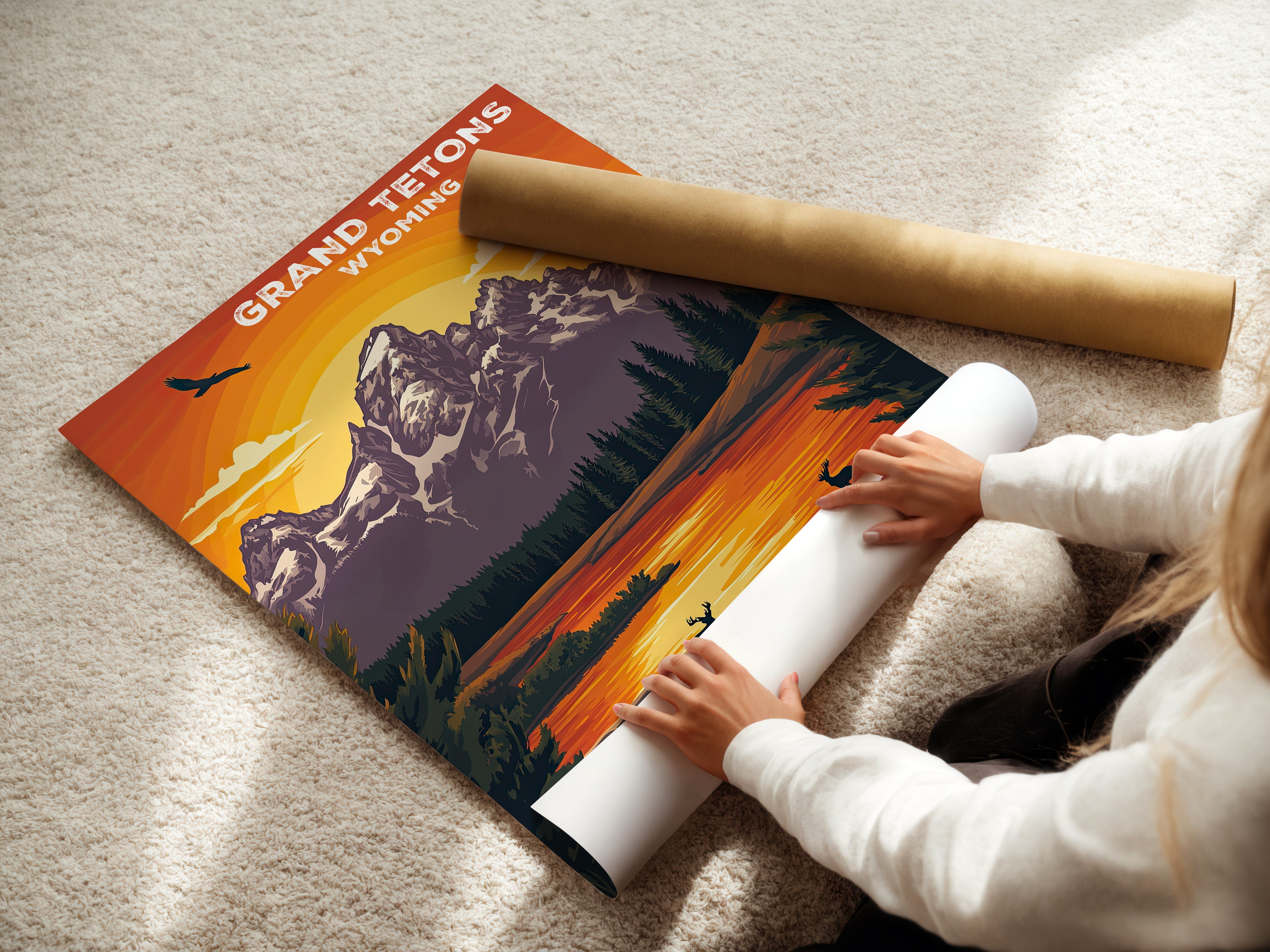 Person unrolling vintage travel poster of Grand Tetons sunset, showcasing mountains and wildlife in Wyoming.